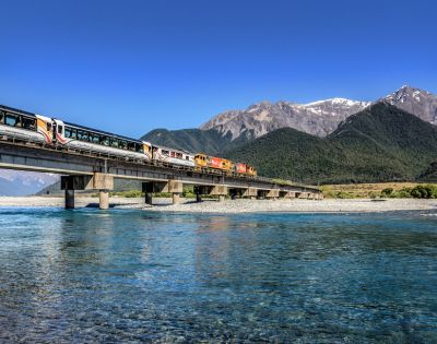 A train travels across a bridge spanning a clear blue river, set against a backdrop of lush green hills and majestic snow-capped mountains under a vibrant blue sky.