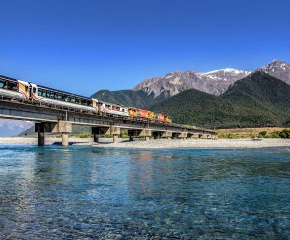A train travels across a bridge spanning a clear blue river, set against a backdrop of lush green hills and majestic snow-capped mountains under a vibrant blue sky.