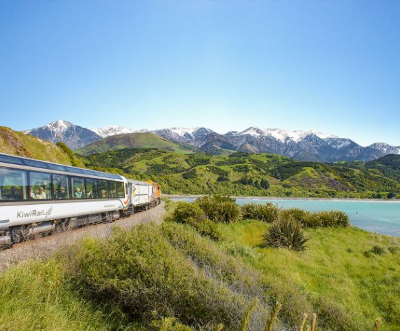 A scenic train travels alongside a turquoise lake, with lush green hills and snow-capped mountains under a clear blue sky in the background.