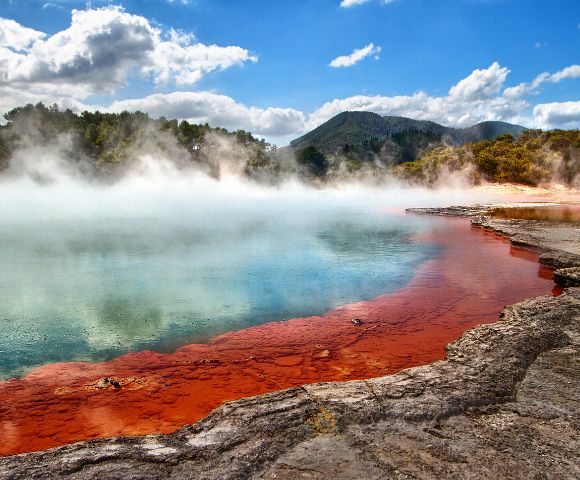 Vibrant hot spring with red, orange, and turquoise hues, surrounded by steam and rocks under a bright blue sky and fluffy clouds. Majestic and serene.
