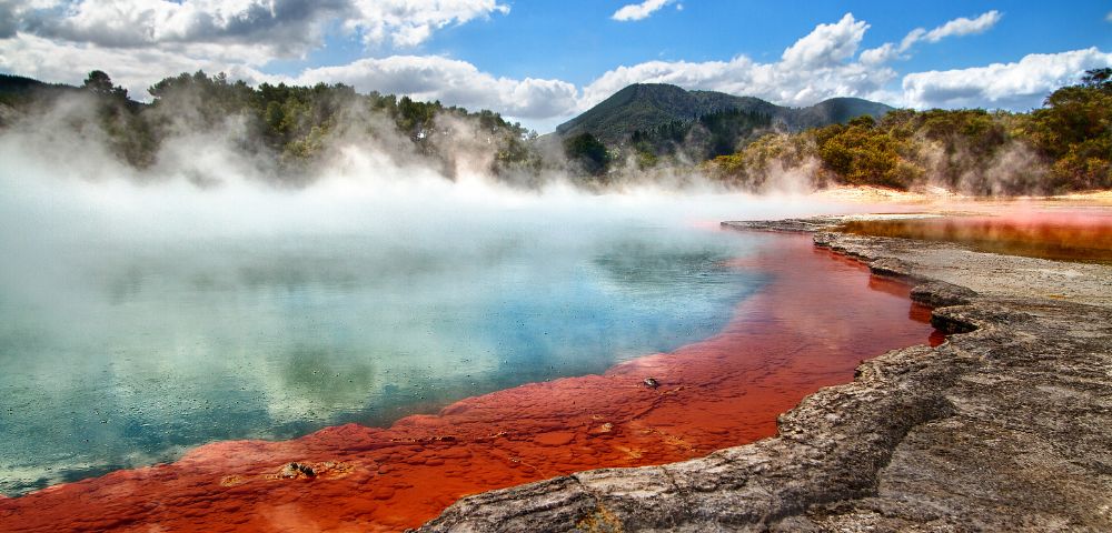 Vibrant hot spring with red, orange, and turquoise hues, surrounded by steam and rocks under a bright blue sky and fluffy clouds. Majestic and serene.