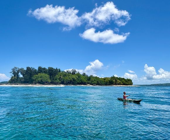 A person paddles a kayak in clear blue water near a small, lush island under a vibrant sky with fluffy clouds, evoking a sense of tranquility and adventure.