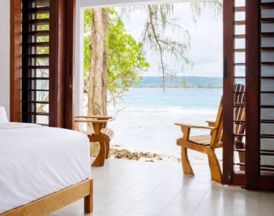 A serene bedroom opens to a beach view with a white bed in the foreground and wooden chairs on a patio overlooking turquoise water and lush greenery.