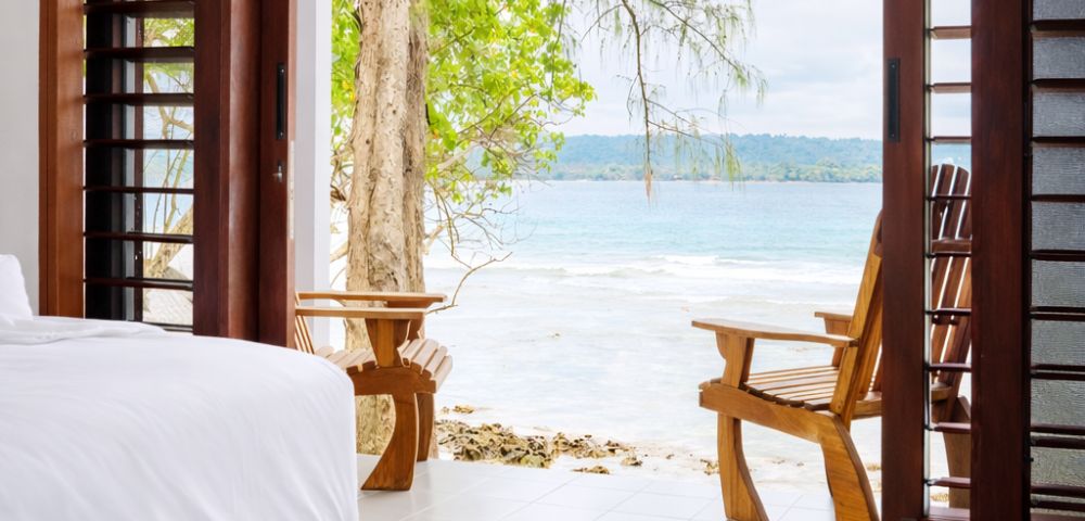A serene bedroom opens to a beach view with a white bed in the foreground and wooden chairs on a patio overlooking turquoise water and lush greenery.