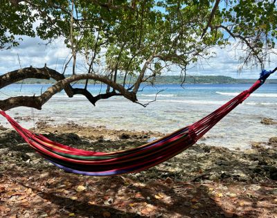 A colorful hammock hangs between tree branches on a rocky beach, overlooking a calm sea under a partly cloudy sky, evoking a sense of tranquility.