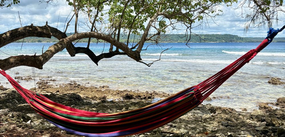 A colorful hammock hangs between tree branches on a rocky beach, overlooking a calm sea under a partly cloudy sky, evoking a sense of tranquility.