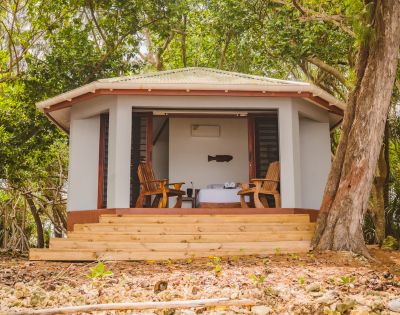 A cozy cabin with a green roof is nestled among lush trees. Two wooden chairs face a table on a porch, evoking relaxation and seclusion.