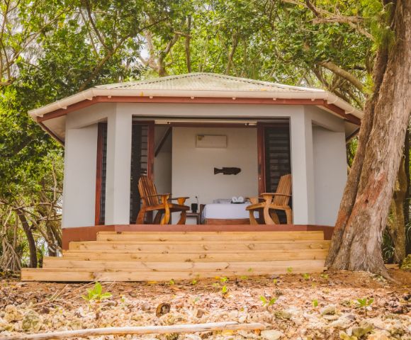 A cozy cabin with a green roof is nestled among lush trees. Two wooden chairs face a table on a porch, evoking relaxation and seclusion.