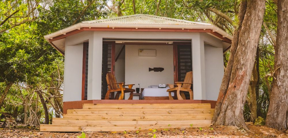A cozy cabin with a green roof is nestled among lush trees. Two wooden chairs face a table on a porch, evoking relaxation and seclusion.