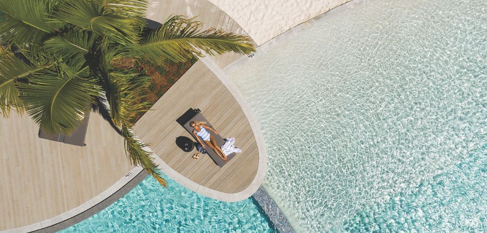 Aerial view of a woman lounging on a wooden deck by a clear pool and sandy beach, surrounded by tropical plants.