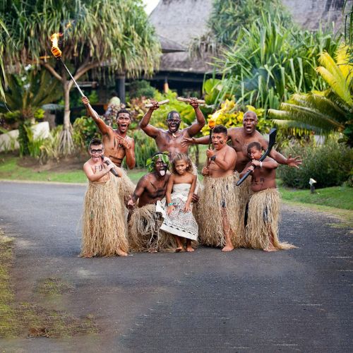 A group of people in traditional attire pose joyfully on a paved path, with grass skirts, face paint, and props. Tropical greenery surrounds them.