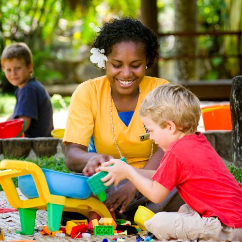 A woman in a yellow shirt, smiling and playing with blocks with a young boy wearing a red shirt. They are outdoors, sitting on grass, with a joyful atmosphere.