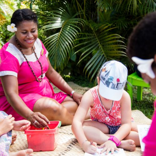 A woman in a pink dress and flower hair accessory smiles while sitting with children drawing outdoors. Lush green plants surround the cheerful scene.
