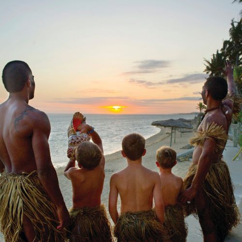 Five people in traditional grass skirts watch a vibrant sunset over the ocean. The scene is calm and reflective, with warm hues and palm trees.