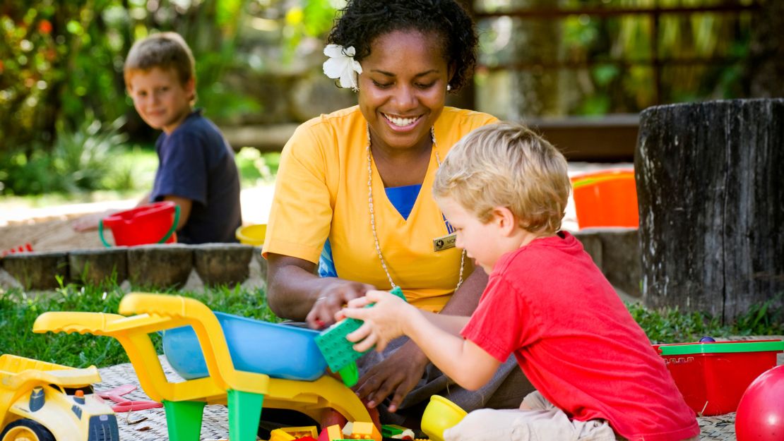 A woman in a yellow shirt, smiling and playing with blocks with a young boy wearing a red shirt. They are outdoors, sitting on grass, with a joyful atmosphere.