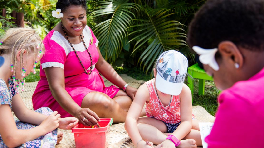 A woman in a pink dress and flower hair accessory smiles while sitting with children drawing outdoors. Lush green plants surround the cheerful scene.