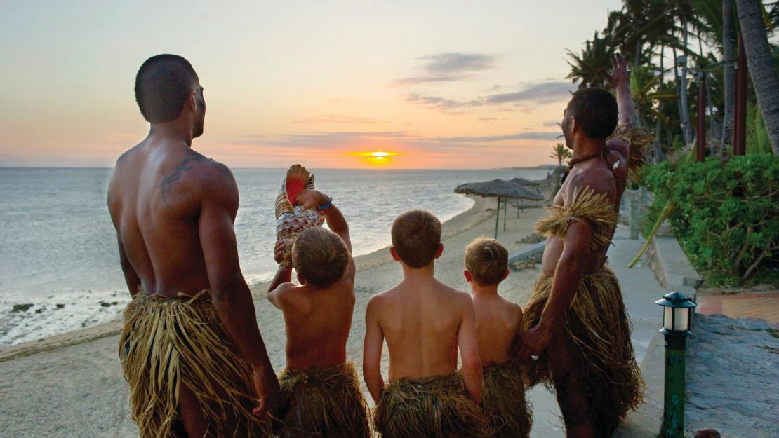 Five people in traditional grass skirts watch a vibrant sunset over the ocean. The scene is calm and reflective, with warm hues and palm trees.