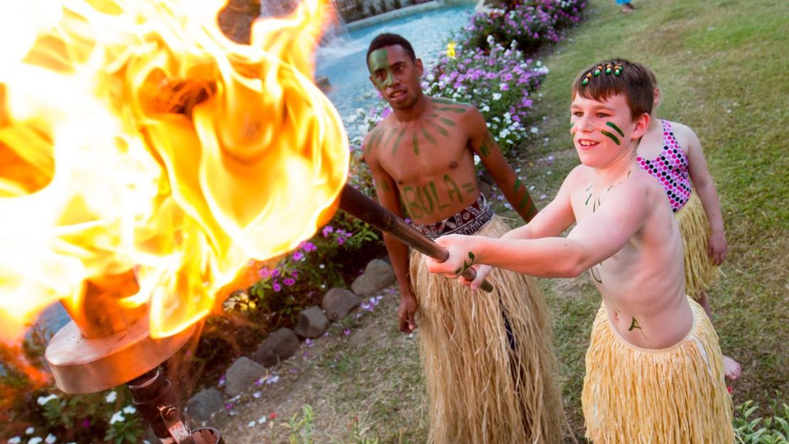 Bare-chested boys in grass skirts light a torch, surrounded by lush greenery and vibrant flowers. The scene feels festive and cultural.