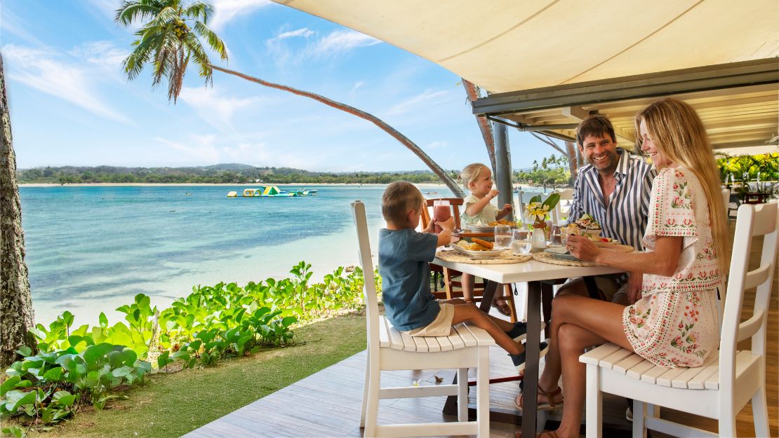 Family enjoying a meal at a beachside restaurant, overlooking turquoise waters and distant hills. The atmosphere is relaxed and joyful.