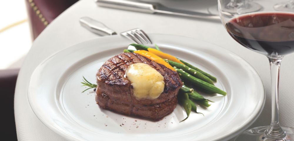A perfectly cooked steak topped with butter, served with green beans and orange slices, alongside a glass of red wine on a white tablecloth.