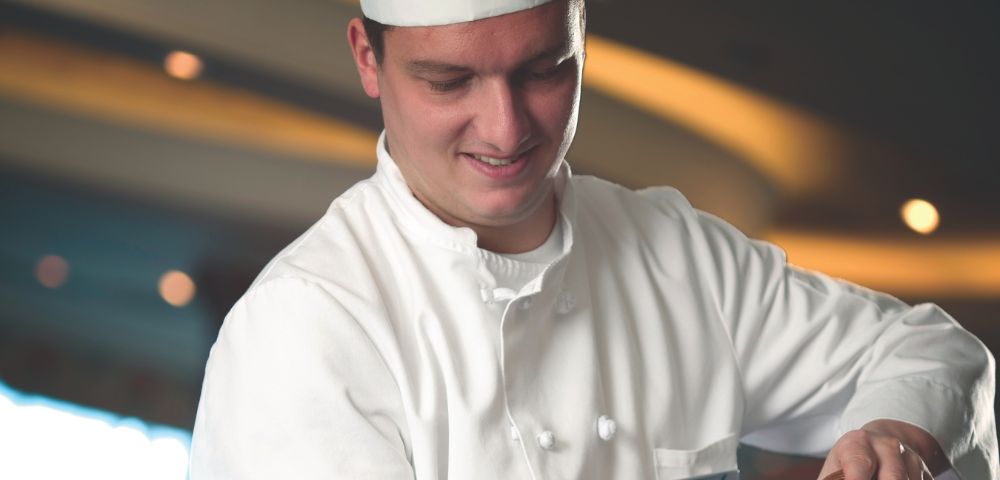 A chef in a white uniform prepares a dish, with soft lighting and a blurred background creating an inviting kitchen atmosphere.