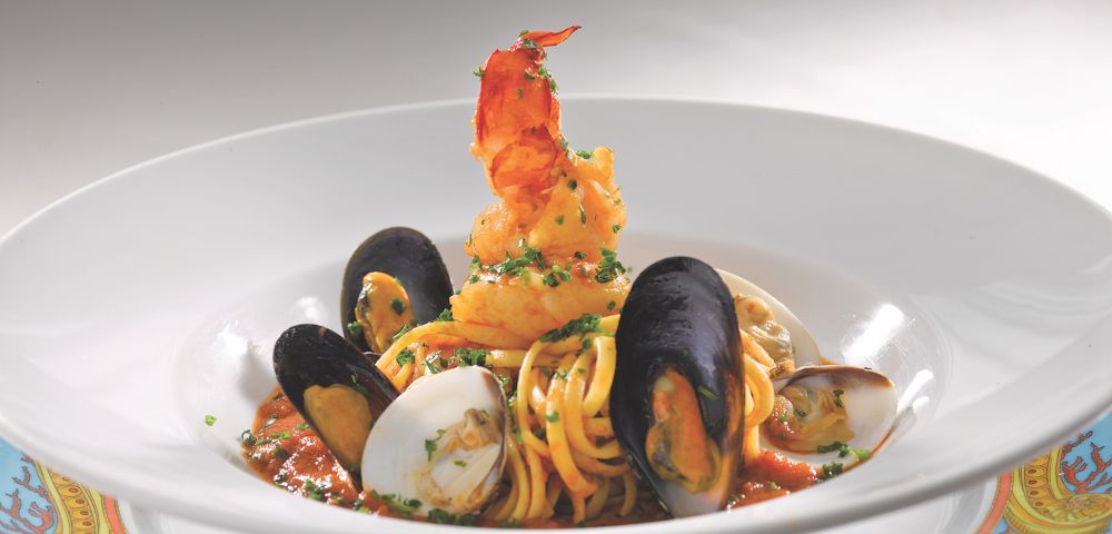 A gourmet seafood pasta dish featuring linguine, mussels, clams, and a large shrimp, garnished with fresh herbs in a savory sauce.
