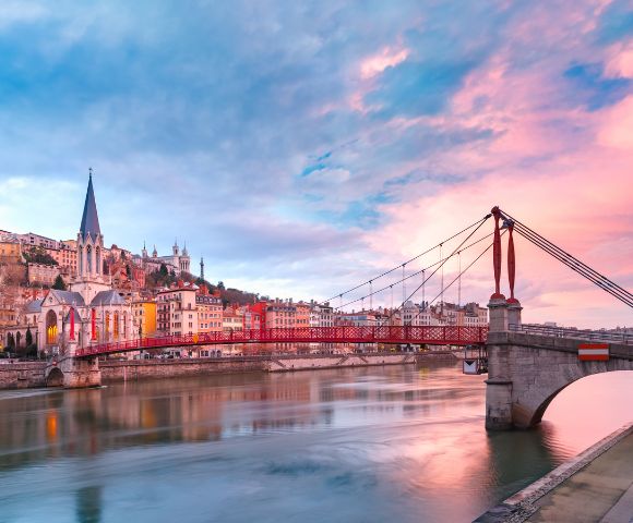 A vibrant sunset over a cityscape with a red suspension bridge spanning a calm river. Colorful buildings and a church with a tall spire line the riverbank.
