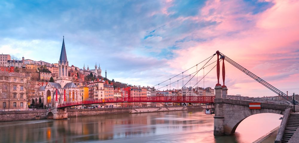 A vibrant sunset over a cityscape with a red suspension bridge spanning a calm river. Colorful buildings and a church with a tall spire line the riverbank.