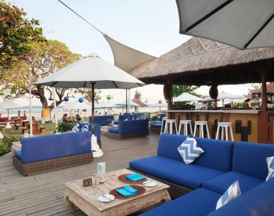 Outdoor beachside patio with blue cushioned sofas, white umbrellas, and a thatched bar. Atmosphere is relaxed and inviting with ocean views.