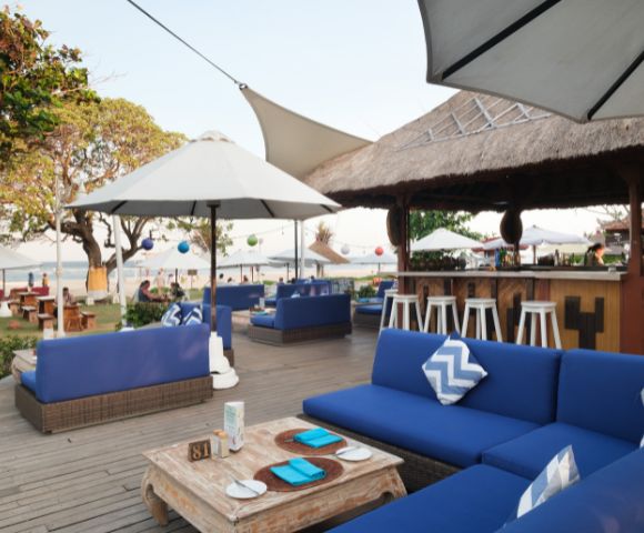 Outdoor beachside patio with blue cushioned sofas, white umbrellas, and a thatched bar. Atmosphere is relaxed and inviting with ocean views.