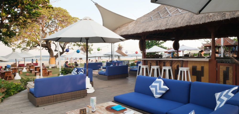 Outdoor beachside patio with blue cushioned sofas, white umbrellas, and a thatched bar. Atmosphere is relaxed and inviting with ocean views.