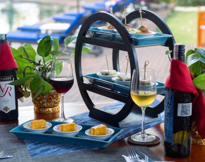 A tiered tray with appetizers, wine glasses, and bottles on a table by a pool. The scene conveys a relaxed, elegant outdoor dining atmosphere.