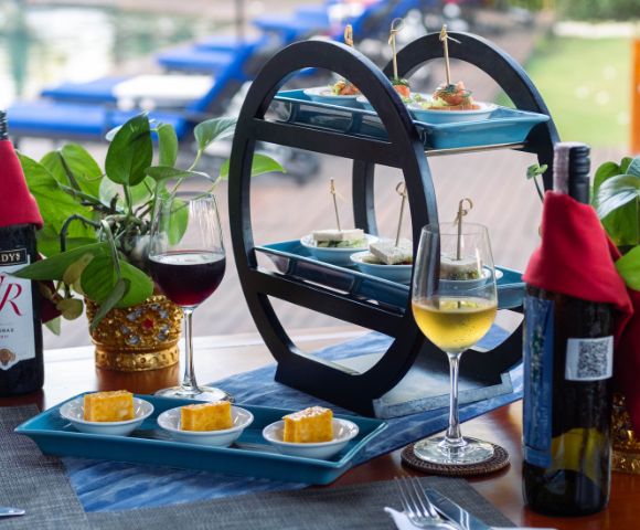 A tiered tray with appetizers, wine glasses, and bottles on a table by a pool. The scene conveys a relaxed, elegant outdoor dining atmosphere.