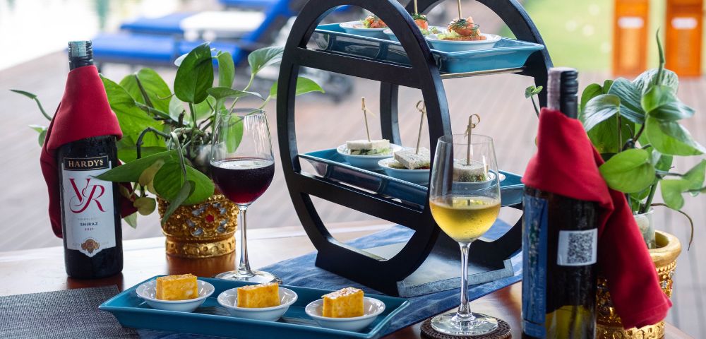 A tiered tray with appetizers, wine glasses, and bottles on a table by a pool. The scene conveys a relaxed, elegant outdoor dining atmosphere.