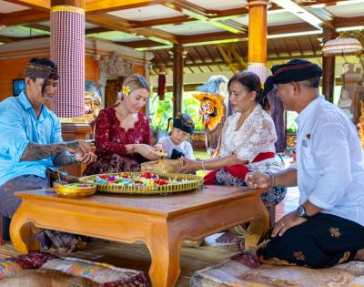 A group of people in traditional attire, including a child, sits on a wooden floor carving wooden masks, conveying a joyful and cultural atmosphere.