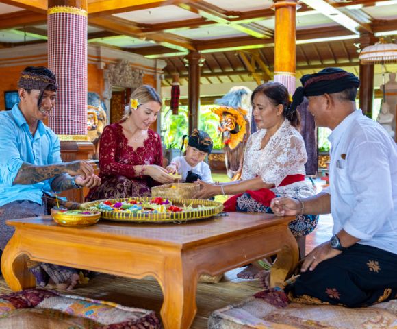 A group of people in traditional attire, including a child, sits on a wooden floor carving wooden masks, conveying a joyful and cultural atmosphere.