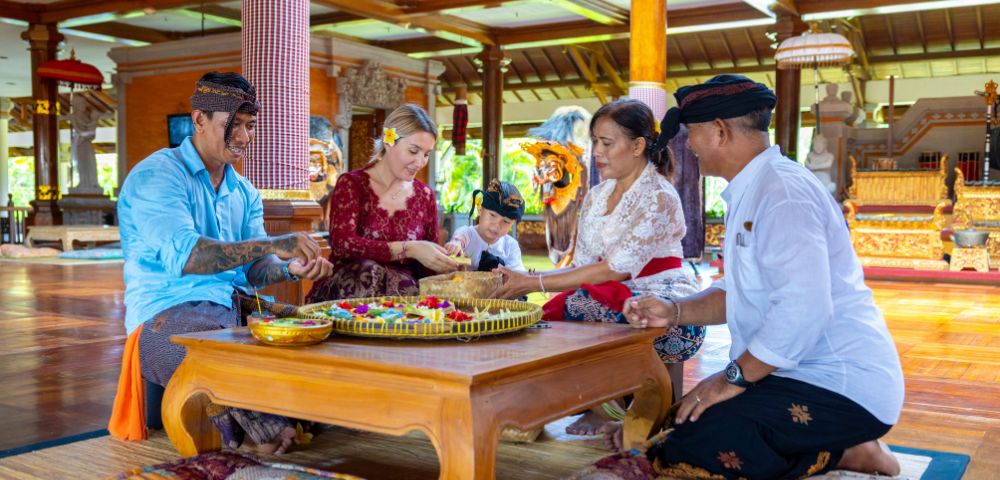 A group of people in traditional attire, including a child, sits on a wooden floor carving wooden masks, conveying a joyful and cultural atmosphere.