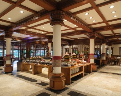 Spacious restaurant interior with wooden beams, columns, and buffet tables filled with dishes. Warm lighting creates a welcoming, elegant atmosphere.