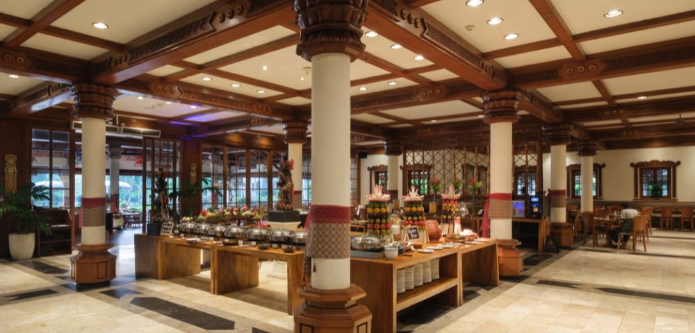 Spacious restaurant interior with wooden beams, columns, and buffet tables filled with dishes. Warm lighting creates a welcoming, elegant atmosphere.