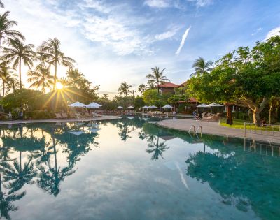 Sunset view of a tranquil pool with palm trees and lounge chairs. The sun casts reflections on the water, creating a peaceful, tropical atmosphere.