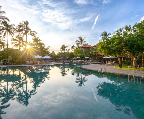 Sunset view of a tranquil pool with palm trees and lounge chairs. The sun casts reflections on the water, creating a peaceful, tropical atmosphere.