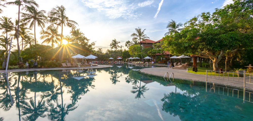 Sunset view of a tranquil pool with palm trees and lounge chairs. The sun casts reflections on the water, creating a peaceful, tropical atmosphere.