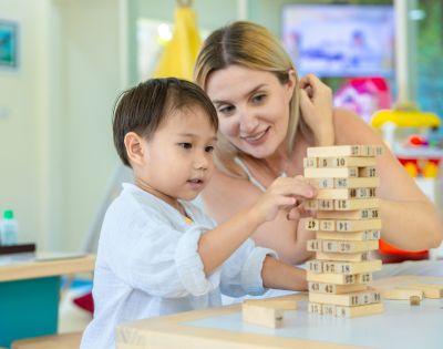 A child and an adult play with a wooden block tower in a brightly lit room. Both are focused and smiling, suggesting engagement and learning.