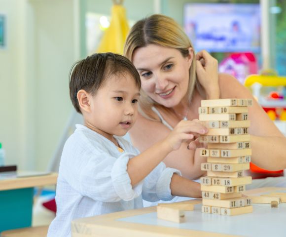 A child and an adult play with a wooden block tower in a brightly lit room. Both are focused and smiling, suggesting engagement and learning.