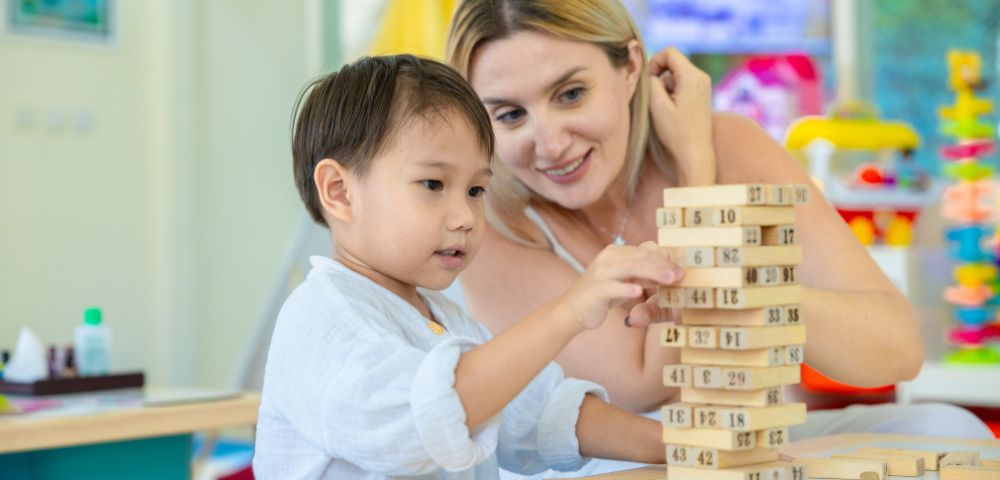 A child and an adult play with a wooden block tower in a brightly lit room. Both are focused and smiling, suggesting engagement and learning.