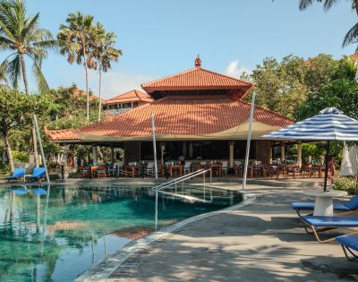 A serene tropical resort poolside scene with palm trees and a traditional building. Cozy blue loungers and striped umbrellas enhance the relaxed ambiance.