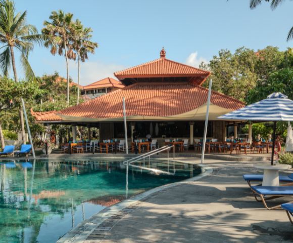 A serene tropical resort poolside scene with palm trees and a traditional building. Cozy blue loungers and striped umbrellas enhance the relaxed ambiance.