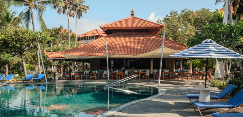 A serene tropical resort poolside scene with palm trees and a traditional building. Cozy blue loungers and striped umbrellas enhance the relaxed ambiance.
