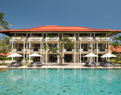 Three-story resort with red-tiled roof, balconies, and white umbrellas. A clear blue pool in the foreground, palm trees, and bright sky create a tranquil atmosphere.