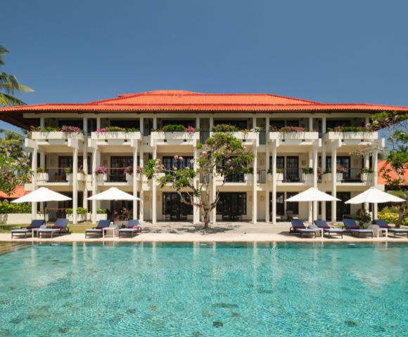 Three-story resort with red-tiled roof, balconies, and white umbrellas. A clear blue pool in the foreground, palm trees, and bright sky create a tranquil atmosphere.